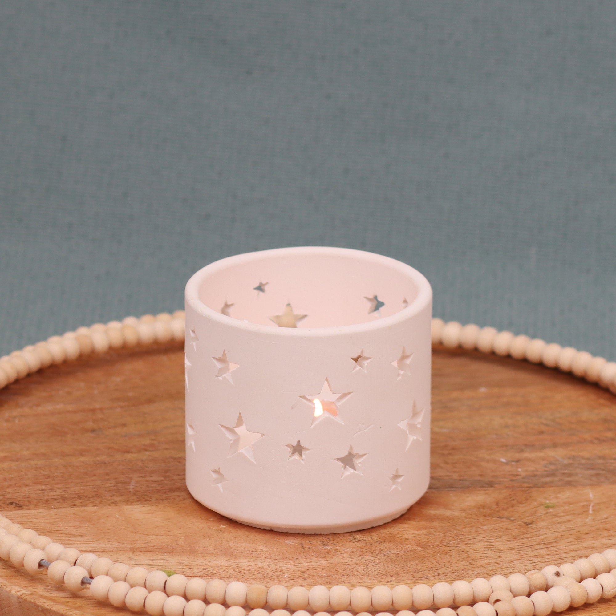 Teelicht-Windspiel-leuchtend-Gießobjekt aus weißen Gips mit Sternenausstanzungen -auf Holzplatte mit Holzperlenkranz vor türkisfarbenem Hintergrund